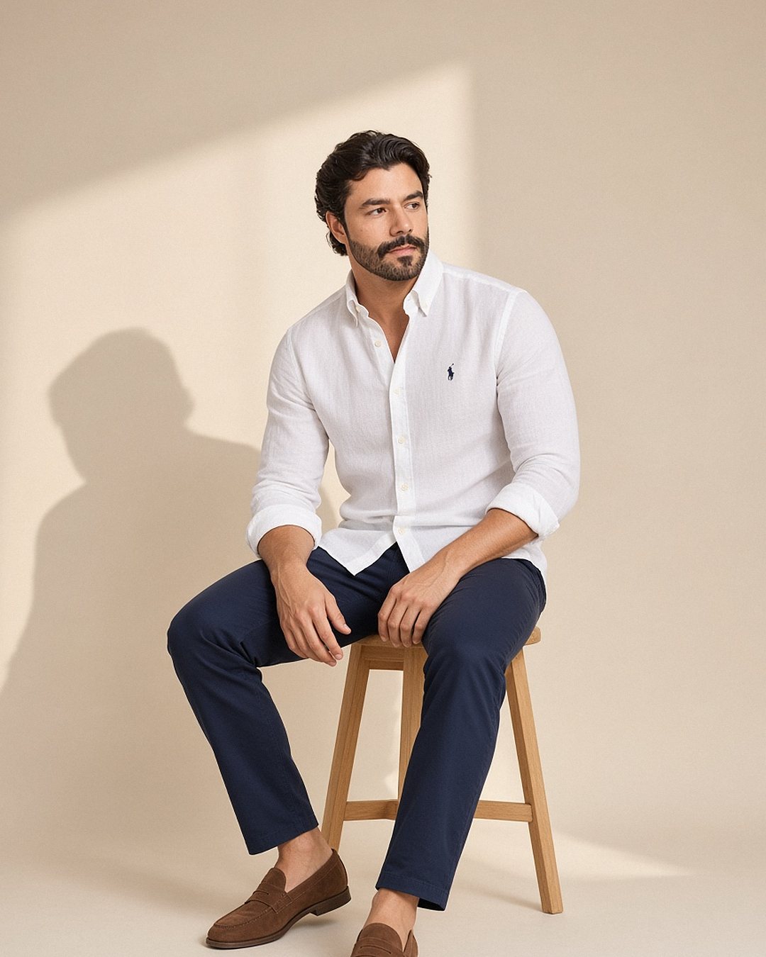 Homem sentado em banco de madeira usando camisa de linho Ralph Lauren branca de mangas dobradas com calça chino azul-marinho e mocassim marrom, em estúdio bege com luz natural de janela