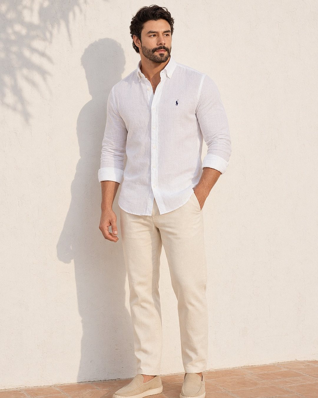 Homem com camisa de linho Ralph Lauren branca de mangas dobradas e dois botões abertos, por fora da calça de linho creme, com mocassim bege, em parede caiada com sombra de folhagem sob o sol da tarde