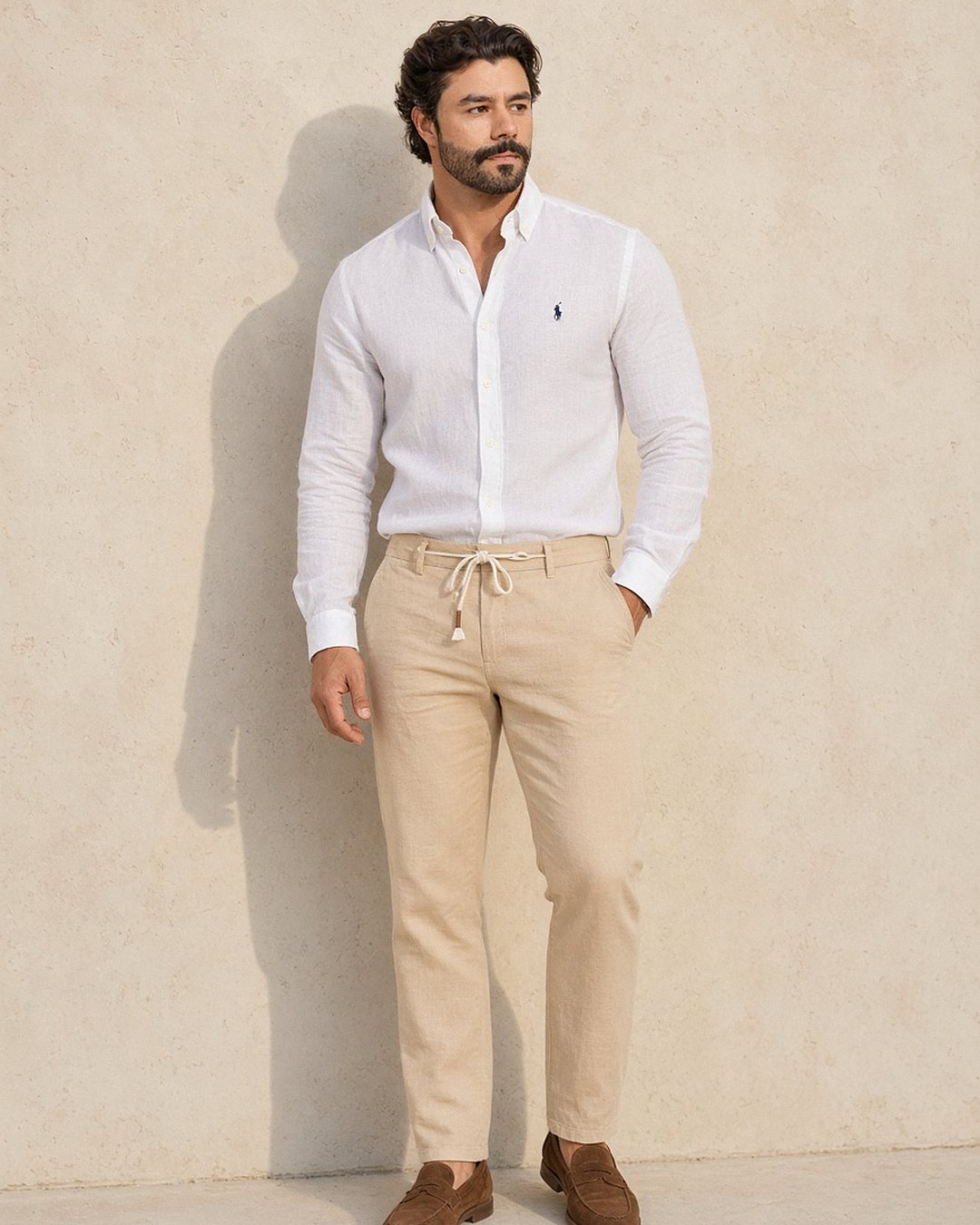 Homem com camisa de linho Ralph Lauren branca por dentro da calça de linho areia com cordão, e mocassim de camurça marrom, em frente a parede clara iluminada pela luz quente do fim de tarde