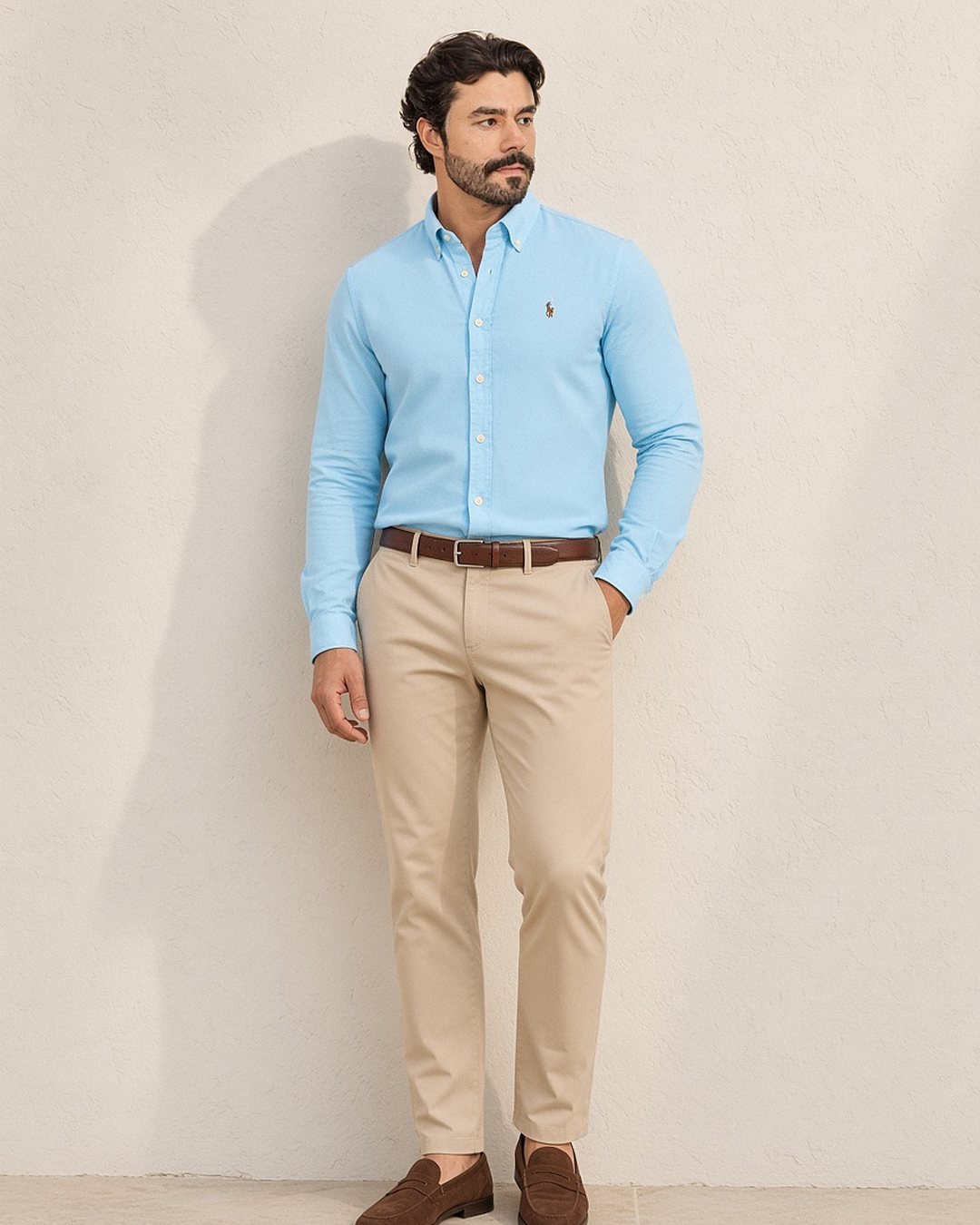 Homem com camisa oxford Ralph Lauren azul-claro de gola button-down por dentro da calça chino bege, com cinto de couro marrom e mocassim de camurça, em frente a parede de cal clara