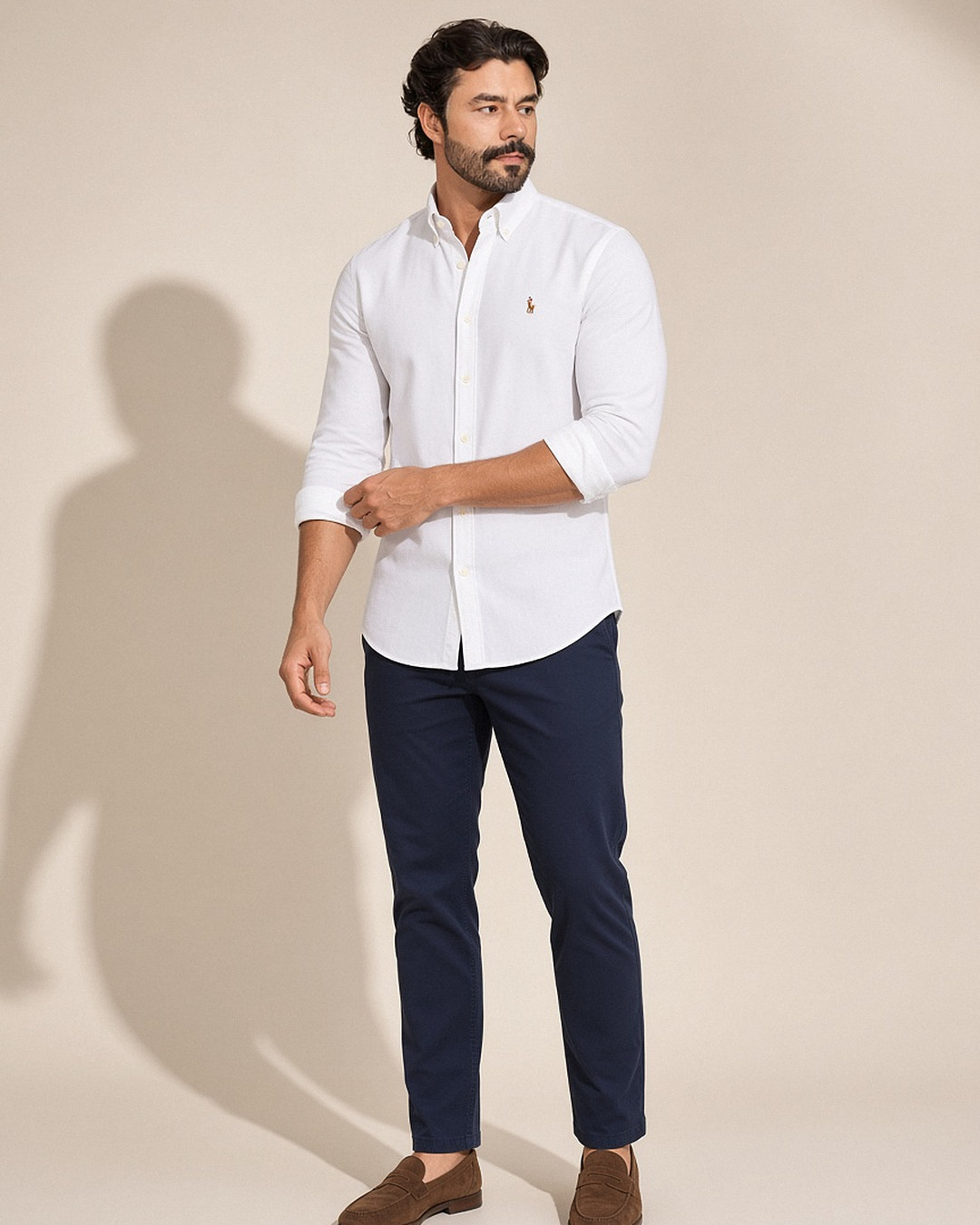 Homem com camisa oxford Ralph Lauren branca de gola button-down com as mangas dobradas, usada por fora da calça chino azul-marinho e com mocassim marrom, em estúdio com luz natural