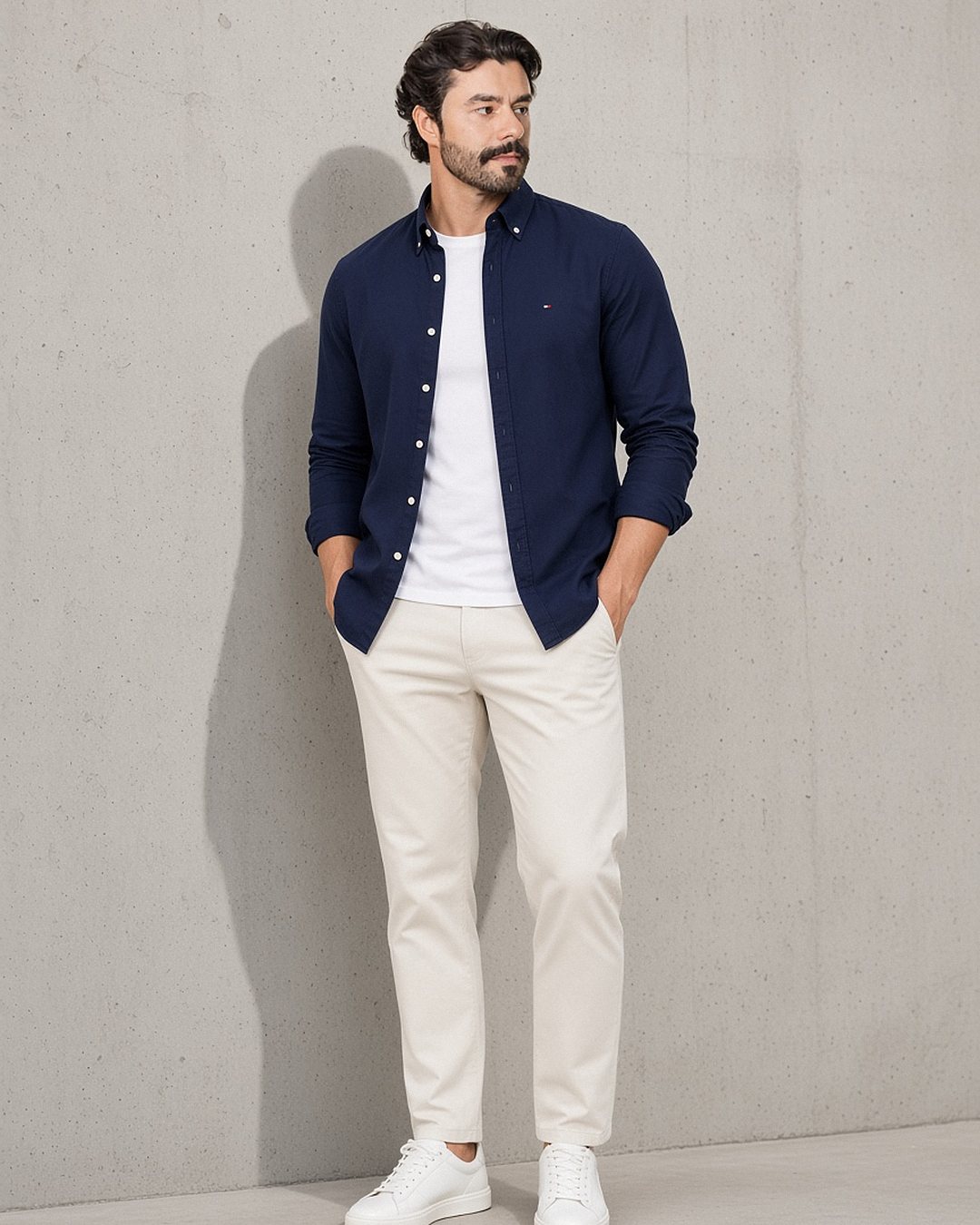Homem com camisa oxford Tommy Hilfiger azul-marinho usada aberta sobre camiseta branca, com calça chino off-white e tênis branco, encostado em parede de concreto aparente