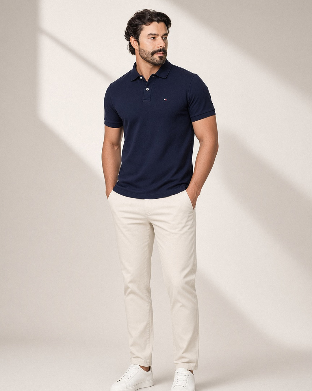 Homem com camisa polo Tommy Hilfiger Custom Fit azul-marinho de piquet com botões brancos e bandeira bordada no peito, usada com calça chino off-white e tênis branco em estúdio com luz natural