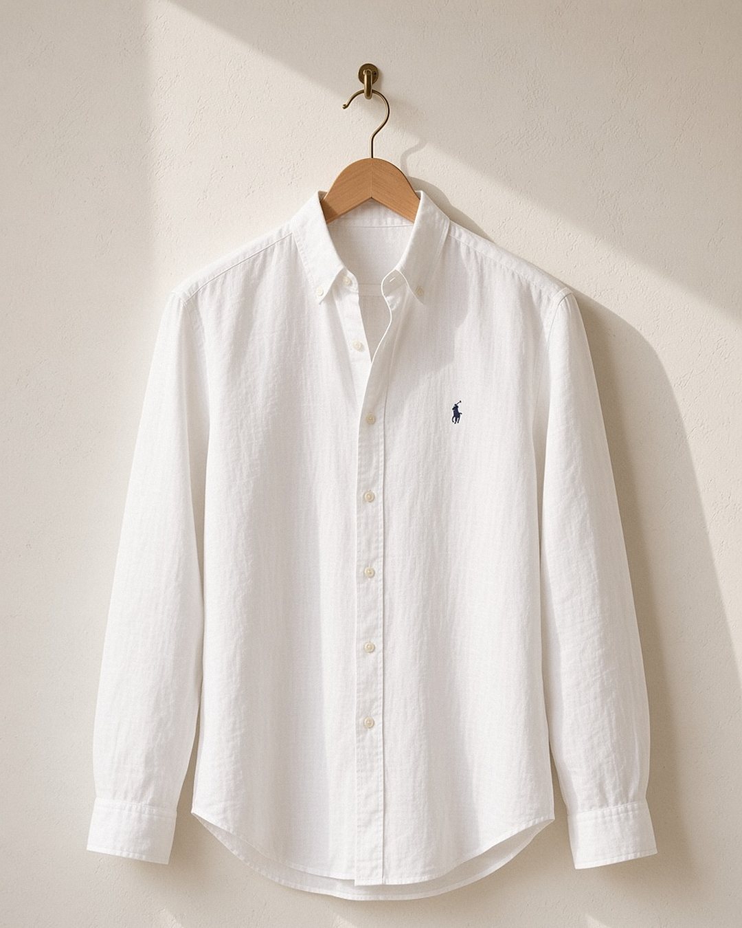 Camisa de linho Ralph Lauren branca pendurada em cabide de madeira contra parede de gesso clara, secando à sombra com luz natural suave